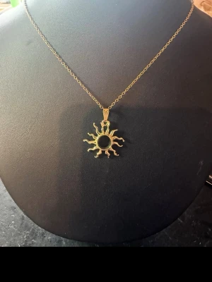 Guldigt solhalsband med svart sten - Unikt halsband med ett solformat hänge i guldfärgad metall. Kedjan är tunn och elegant, och hänget har detaljerade solstrålar som ger en cool och bohemisk vibe. Perfekt för dig som vill sticka ut med accessoarer som har karaktär.