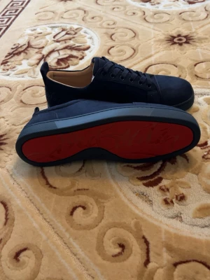 Mörkblåa Louboutin sneakers med röd sula - Snygga svarta sneakers från Christian Louboutin med klassisk röd sula och diskret logga undertill. Skorna har lågt skaft, svarta skosnören och ovandel i mocka. Perfekta för dig som vill ha en exklusiv och stilren look.