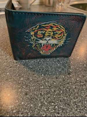 Ed Hardy Plånbok med tiger och dödskallemotiv - Cool plånbok i mörkgrönt konstläder med färgstarkt tryck av en tiger på ena sidan och dödskalle med texten 'Death or Glory' samt stjärnor på andra sidan. Plånboken har en klassisk vikbar form och detaljerade illustrationer i tatueringsstil.