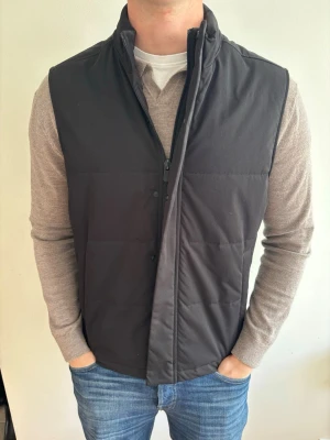 Svart dunväst med ståkrage - Storlek L. Stilren svart dunväst med ståkrage och dragkedja framtill. Västen har en enkel, quiltad design och är perfekt att bära över en tröja för extra värme. Passar till jeans eller andra avslappnade outfits.