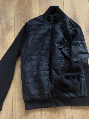 J Lindberg cardigan - Svart cardiganjacka med quiltad front och stickade ärmar samt rygg. Jackan har hög krage, hel dragkedja och fickor med dragkedja. Materialet är en kombination av stickat tyg och quiltad nylon. 