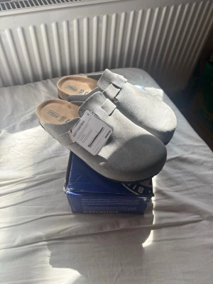Grå mockatofflor Birkenstock Boston  - Säljer ett par snygga grå tofflor från Birkenstock i mjuk mocka. Modellen har en klassisk design med justerbart spänne i metall och bekväm innersula. Perfekta för dig som vill ha både stil och komfort hemma eller på språng. Helt nya aldrig använda endast testade inomhus! Hör av er vid eventuella frågor!