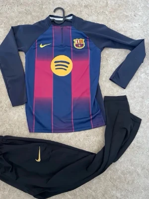 FC Barcelona träningsset Nike - Snyggt träningsset från Nike med FC Barcelona-tema. Tröjan har lång ärm, halv dragkedja, blå och vinröda ränder samt klubbmärke och Spotify-logga. Byxorna är svarta med gul Nike-logga. Perfekt för fotbollsträning eller sportiga dagar.