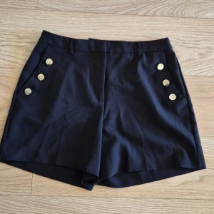 Svarta shorts med guldknappar - Stilrena svarta shorts med hög midja och dekorativa guldknappar framtill. Klassisk passform med bälteshällor och fickor. Perfekta för en trendig och chic look.