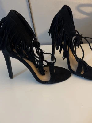 Svarta pumps med fransar och snörning - Säljer ett par svarta pumps med höga klackar, fransdetaljer och snörning runt ankeln. Skorna har öppen tå och är tillverkade i mockaimitation som ger en lyxig känsla. Perfekta för dig som vill sticka ut med en trendig och edgy stil. Tror dom är från hm kommer ej ihåg 