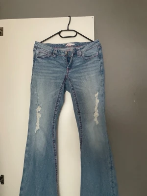 Ljusblå bootcut jeans med brodyr - Säljer ett par ljusblå bootcut jeans med slitningar framtill och rosa broderade detaljer på bakfickorna. Jeansen har klassisk femficksmodell och är tillverkade i denim med lätt stretch. Perfekta för en avslappnad och trendig look.
