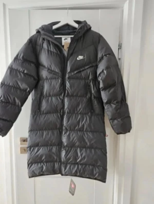 Svart lång dunjacka från Nike - Snygg svart lång dunjacka från Nike med huva och dragkedja framtill. Jackan har två sidofickor med dragkedja och diskret Nike-logga på bröstet. Perfekt för kalla vinterdagar och håller dig varm med sin vadderade design. Sitter som S jätte fin jacka