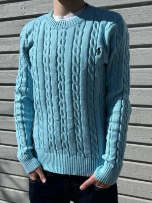 Ljusblå kabelstickad tröja Polo Ralph Lauren - Ljusblå Ralph Lauren kabelstickad tröja med blå Ralph Lauren-logga på bröstet. Helt ny med tags kvar. Storlek S och sitter clean i passformen. Lätt att styla med jeans, cargos eller över en skjorta.