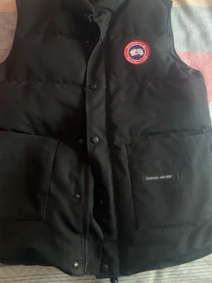 Svart dunväst från Canada Goose - Svart dunväst från Canada Goose med klassisk logga på bröstet och två stora fickor framtill. Västen har dragkedja och tryckknappar, samt är fodrad med dun för extra värme. Perfekt för lager-på-lager och streetstyle.