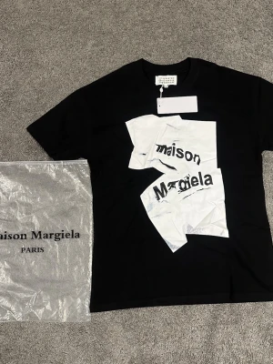 Maison Margiela svart t-shirt - Cool svart t-shirt från Maison Margiela med unik vit grafisk print och logga framtill. Klassisk passform med rund hals och korta ärmar. Perfekt för dig som gillar statement-plagg och streetstyle.
