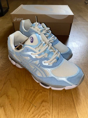 Asics GEL-NYC - Snygga ASICS sneakers i en fräsch kombination av ljusblått och beige. Skorna har meshpaneler för bra ventilation, detaljer i mocka och syntet samt vita snören. Ikonisk ASICS-logga på sidan och tungan. Perfekta för dig som gillar sportig och trendig stil.