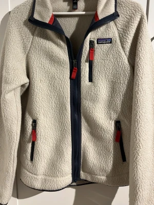 Vit fleecejacka från Patagonia - Säljer en vit fleecejacka från Patagonia med marinblå dragkedjor och röda detaljer. Jackan har tre fickor med dragkedja och klassisk logga på bröstet. Perfekt för lager-på-lager och chill dagar. Materialet är mjuk och fluffig fleece.  Nypris 1700kr.