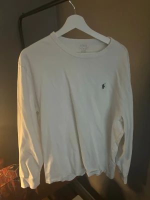 Vit långärmad tröja från Polo Ralph Lauren - Vit långärmad tröja från Polo Ralph Lauren med klassisk rund halsringning och den ikoniska blå broderade loggan på bröstet. Tillverkad i mjuk bomull, perfekt för en clean och stilren look.