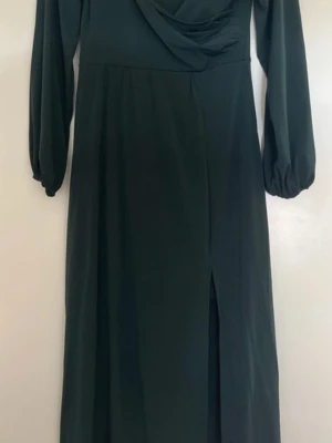 Mörkgrön långklänning med slits - Stilren mörkgrön långklänning med långa ärmar och draperad urringning. Klänningen har en diskret slits framtill och är tillverkad i ett mjukt, lätt material som faller snyggt. Perfekt för dig som gillar en elegant och enkel look.