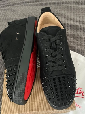 Christian Louboutin svarta spikes sneakers - Svarta sneakers från Christian Louboutin med mattsvarta nitar på tån och sidorna. Skorna har klassisk röd sula, svarta skosnören och ovandel i mocka. Insidan är ljusbeige och designen är riktigt edgy och lyxig.