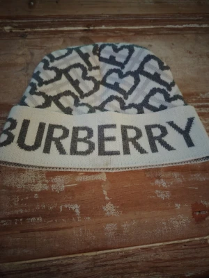 Vit och grå Burberry mössa - Snygg stickad mössa från Burberry i vitt och grått med stort logotyptryck framtill och mönster av sammankopplade B:n. Perfekt accessoar för att lyfta din stil under kalla dagar.