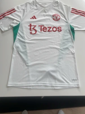 Manchester United bortatröja Adidas - Snygg vit Manchester United bortatröja från Adidas med röda detaljer och klubbmärke på bröstet. Tröjan har gröna inslag på sidorna, röda ränder på axlarna och Tezos som sponsortryck. Tillverkad i lätt och ventilerande material, perfekt för fotboll eller träning.