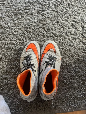 Nike grå-orange fotbollsskor med socka - Säljer ett par gråa Nike fotbollsskor med högt skaft i strumpmodell och starka orange detaljer. Skorna har svarta snören och en cool grafisk design på sidorna. Perfekta för dig som vill sticka ut på planen och ha bra grepp och komfort.