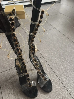 Svarta sandaler med strassdetaljer - Säljer ett par svarta sandaler från Giuseppe Zanotti med transparenta remmar och höga klackar. Sandalerna har dekorativa strassdetaljer i guld och svart längs remmarna som går upp längs benet. Perfekta för dig som vill sticka ut med en lyxig och trendig look.