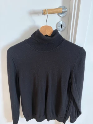Svart polotröja i stickat material - En svart stickad polotröja med lång ärm och ribbade muddar vid ärmslut och nederkant. Tröjan har en klassisk och enkel design, perfekt för lager-på-lager och passar till många olika stilar.