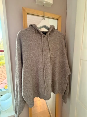 Grå stickad hoodie med huva - Mysig grå stickad hoodie med huva och dragsko. Oversized passform och långa ärmar, perfekt för en avslappnad stil. Materialet är mjukt och ger en chill vibe. Passar perfekt till jeans eller mjukisbyxor.