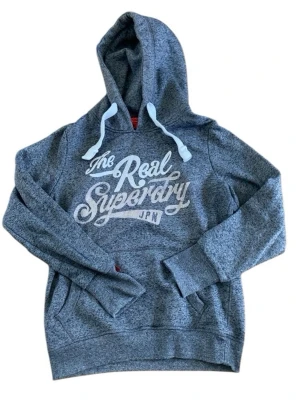 Grå Superdry hoodie med tryck - Mysig grå hoodie från Superdry med vit text på bröstet och klassiska dragsnören i huvan. Hoodien har känguruficka framtill och ribbade muddar vid ärmslut och nederkant. Perfekt för en avslappnad och cool stil.