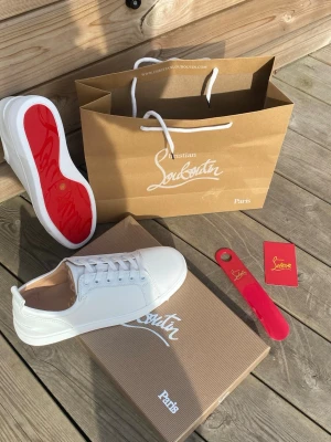 Vita sneakers från Christian Louboutin - Snygga vita sneakers från Christian Louboutin med klassisk röd sula och stilren design. Skorna har vita skosnören, diskreta sömmar och är tillverkade i slätt läder. Perfekta för dig som vill ha en clean look med exklusiv känsla.