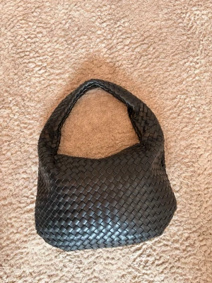 Svart flätad handväska i läder - Snygg svart handväska med flätad design och mjuk, rundad form. Väskan har ett bredare axelband och är tillverkad i läder. Perfekt för dig som gillar stilrena accessoarer med en twist.