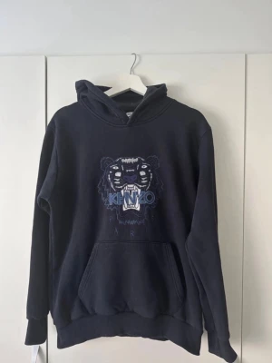 Mörkblå hoodie med tigertryck - Snygg mörkblå hoodie från med det ikoniska tigerbroderiet på bröstet. Hoodien har huva och en stor magficka. Perfekt för dig som gillar streetwear och vill sticka ut med en klassisk design. Pris kan diskuteras
