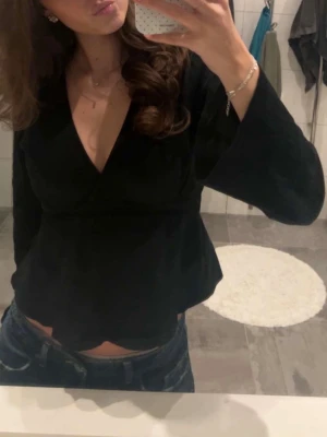 Svart omlottopp med knyt i midjan - Svart omlottopp med v-ringning och långa ärmar. Toppen har knyt i midjan som ger en snygg passform och är tillverkad i ett mjukt tyg. Perfekt att styla med jeans eller kjol för en trendig look.