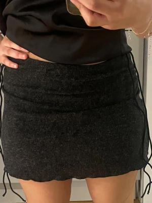 Mörkgrå kortkjol med snörning - Säljer en mörkgrå kortkjol med snörning i sidorna. Kjolen har en tight passform och är gjord i ett mjukt, stretchigt material som känns bekvämt. Perfekt att styla med en crop top eller oversized tröja.