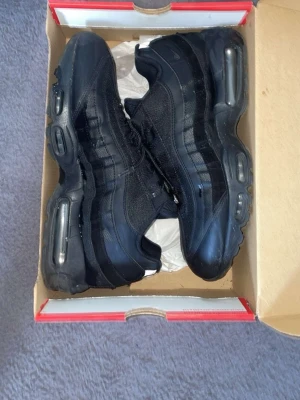 Nike Air Max 95 Triple Black sneakers - Snygga Nike Air Max 95 Triple Black sneakers med ikonisk vågig design. Skorna är helsvarta med mesh, mocka och läderdetaljer samt synliga Air Max-enheter i sulan för extra komfort. Perfekta för dig som gillar streetwear och sportig stil.