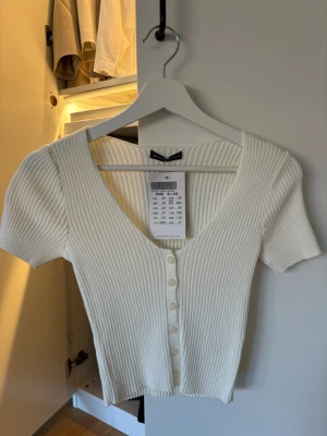 Vit ribbad topp från Brandy Melville - Superfin vit ribbad topp från Brandy Melville med korta ärmar och v-ringning. Toppen har knappar framtill och är croppad, perfekt att styla med jeans eller kjol. Enkel och stilren design som passar till många outfits.