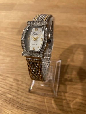 Vintage Etier Quartz Damklocka med Strass - Elegant Etier Quartz damklocka med en rektangulär boett prydd med strass runt urtavlan. Urtavlan är vit med guldfärgade visare och markeringar, samt siffrorna 12, 3, 6 och 9. Klockan har ett tvåfärgat länkarmband i silver- och guldfärgade toner. Baksidan av boetten är märkt med ”STAINLESS STEEL”. Klockan drivs av ett kvartsurverk. Fint skick enligt bilder, behöver bara nytt batteri.  Begagnat värde: < 500 SEK Mitt pris: 189 SEK  Skickas alltid snabbt och spårbart. Skriv vid frågor och se gärna m