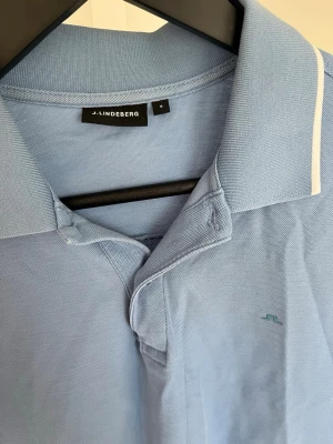Ljusblå pikétröja från J.Lindeberg - Säljer en ljusblå pikétröja från J.Lindeberg med klassisk krage och diskret logga på bröstet. Tröjan har korta ärmar och vit detalj på kragen. Hör gärna av dig vid frågor!