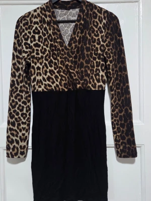 Leopardmönstrad omlottklänning - Snygg kort klänning med leopardmönstrad överdel och svart kjoldel. Omlott-skärning upptill och långa ärmar. Perfekt för dig som vill sticka ut med ett djurmönster och gillar trendiga detaljer.