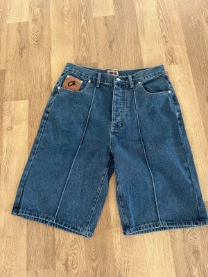 Blå denimshorts från Corteiz  - Snygga blå denimshorts från Corteiz med klassisk femficksdesign och kontrastsömmar. Shortsen har en normal passform och är tillverkade i bomull. Perfekta för en avslappnad streetstil.