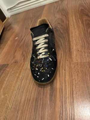 Maison Margiela svart sneakers - Unika svarta sneakers från Maison Margiela med färgstänk i vitt, gult, blått och rött. Skorna har beige sula och vita snören. Tillverkade i läder med klassisk låg siluett och diskret logotyp på plösen. Perfekt för dig som gillar statement-detaljer.