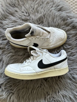 Vita Nike sneakers med svart swoosh - Klassiska vita Nike sneakers med svart swoosh på sidan. Lågt skaft och perforerad tå för extra ventilation. Ovandel i läder och sula i gummi. Perfekta för en clean och sportig look.