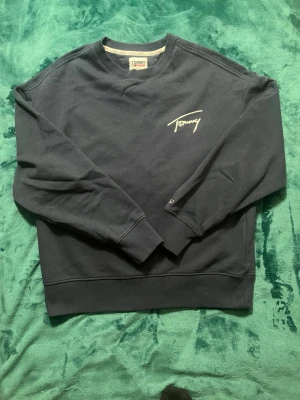Mörkblå sweatshirt Tommy Jeans - Mörkblå sweatshirt från Tommy Jeans med broderad logga i vitt på bröstet. Klassisk rund halsringning och ribbade muddar vid ärmslut och nederkant. Tillverkad i mjuk bomullsblandning för skön känsla.