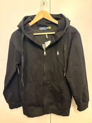 Svart hoodie från Polo Ralph Lauren - Svart hoodie med dragkedja från Polo Ralph Lauren. Klassisk design med huva och dragsnören, två fickor framtill och den ikoniska vita logotypen broderad på bröstet. Tillverkad i mjukt bomullsmaterial som är skönt att bära.