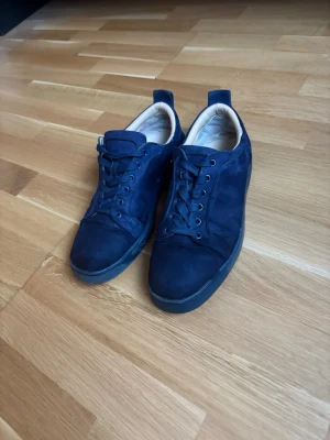 Mörkblå Loubs  - Snygga sneakers i mörkblå mocka med svarta snören och svart sula. Insidan är ljusbeige och yttersulan har en cool röd detalj undertill. Skorna har en klassisk låg profil och stilren design som passar till det mesta. Dem är använda och på sista bilden så ser ni att en knapp lossnat lite men inget som syns så mycket.