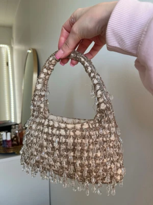 Beige pärlbroderad handväska - Superfin beige handväska med glittrande pärlor och droppformade pärldetaljer över hela väskan. Väskan har en mjuk, rundad form och ett kort handtag, perfekt att bära i handen. Materialet är tyg med pärlbroderi som ger en lyxig och trendig känsla. OBS! Några pärlor har ramlat så det sticker ut lite trådar men inget för synligt. kommer med en kedja och en dustbag. 20 cm bredd, 22 cm lång. Skriv för mer info💗