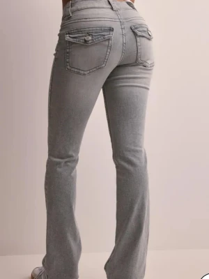 Grå bootcut jeans med fickor - Snygga grå bootcut jeans med klassiska femficksdetaljer och knappar på bakfickorna. Jeansen har låg midja och bootcut . Perfekt för dig som gillar en avslappnad men trendig look.