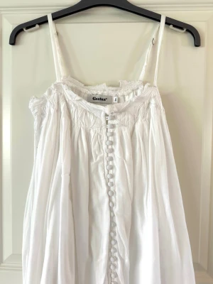 Vit midsommar sommarklänning från Gestuz  - Luftig vit klänning från Gestuz med smala axelband och dekorativa knappar framtill. Klänningen har en romantisk känsla med rynkade detaljer upptill och lätt volang vid halsringningen. Perfekt för varma dagar och enkel att styla.