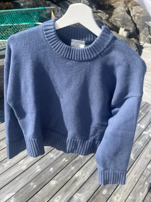 Blå stickad tröja med ribbade detaljer - Säljer en blå stickad tröja med bred ribbad mudd vid hals, ärmslut och nederkant. Tröjan har en avslappnad passform och är perfekt för lager-på-lager. Materialet känns mjukt och bekvämt mot huden.