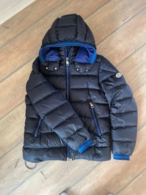 Mörkblå dunjacka från Moncler - Snygg mörkblå dunjacka från Moncler med blå detaljer och huva. Jackan har två dragkedjefickor, ribbade muddar och klassisk Moncler-logga på ärmen. Perfekt för kalla vinterdagar och tillverkad i glansigt material med dunfyllning.