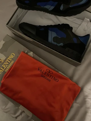 Valentino Garavani blå camo sneakers - Snygga sneakers från Valentino Garavani med blå och svarta camo-detaljer. Skorna har låg profil, svart sula och klassisk snörning. Kommer med originalkartong och röd dustbag. Perfekt för dig som vill sticka ut med exklusiva och trendiga sneakers.