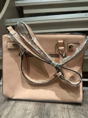 Ljusrosa fyrkantig handväska - Stilren ljusrosa handväska i fyrkantig modell med både handtag och avtagbar axelrem. Väskan har guldfärgade metalldetaljer och svarta kanter. Tillverkad i slätt konstläder och passar perfekt för dig som gillar en clean look.