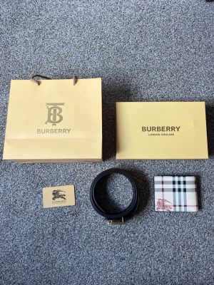 B - bälte och plånbok i klassiskt rutmönster - Snygg plånbok från B i deras ikoniska beigea rutmönster med svarta, vita och röda ränder. Plånboken har en diskret B-logga i hörnet och är tillverkad i tåligt läder. Perfekt för dig som gillar stilrena accessoarer med lyxig känsla. Bältet är 110cm